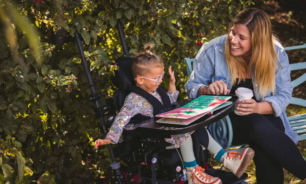 Doris seated in her Hilo wheelchair with the activity tray attached, looking at a book with her mum who is sitting next to her on a bench