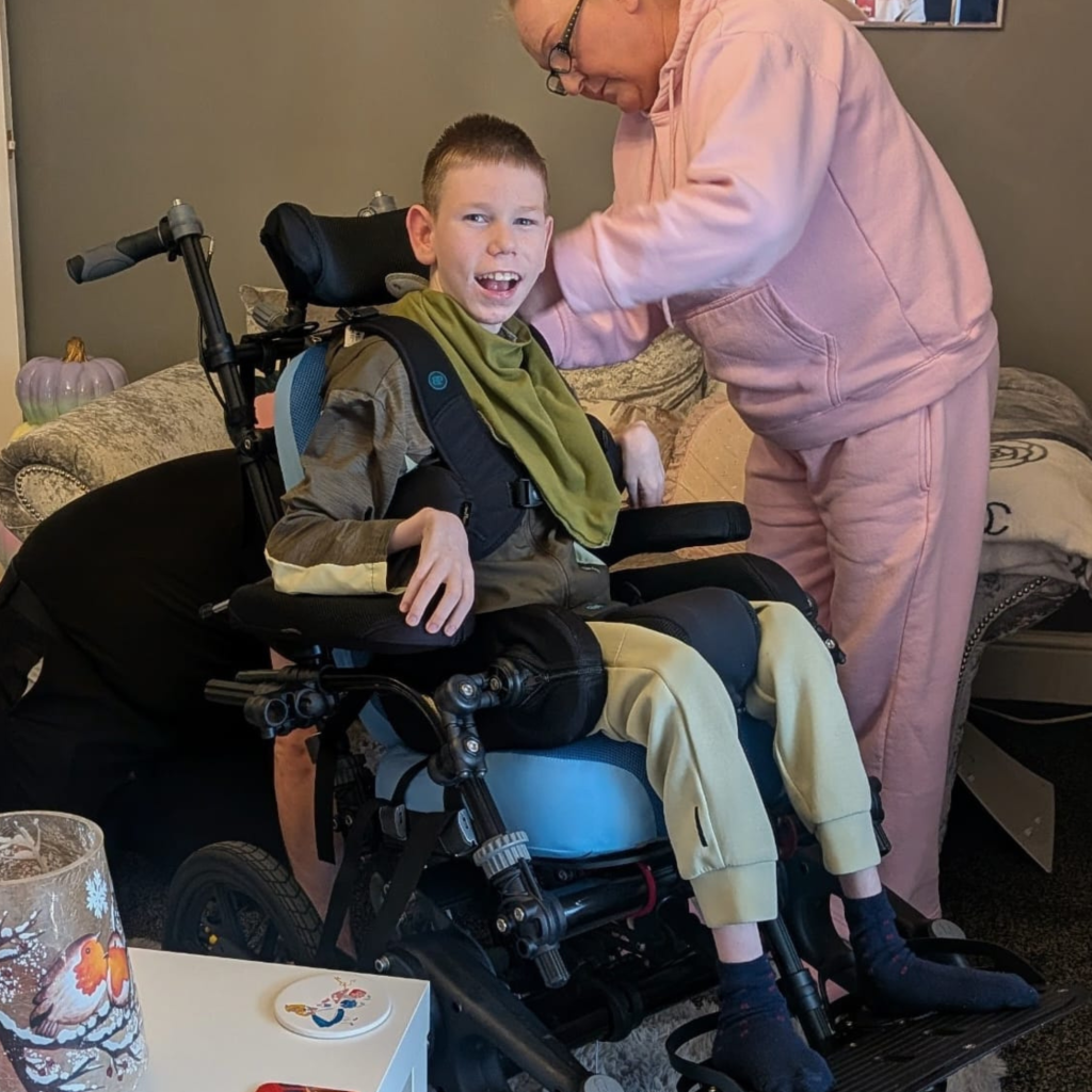 Kian smiling at the camera while seated in his assessment chair as his mum adjusts his seating system